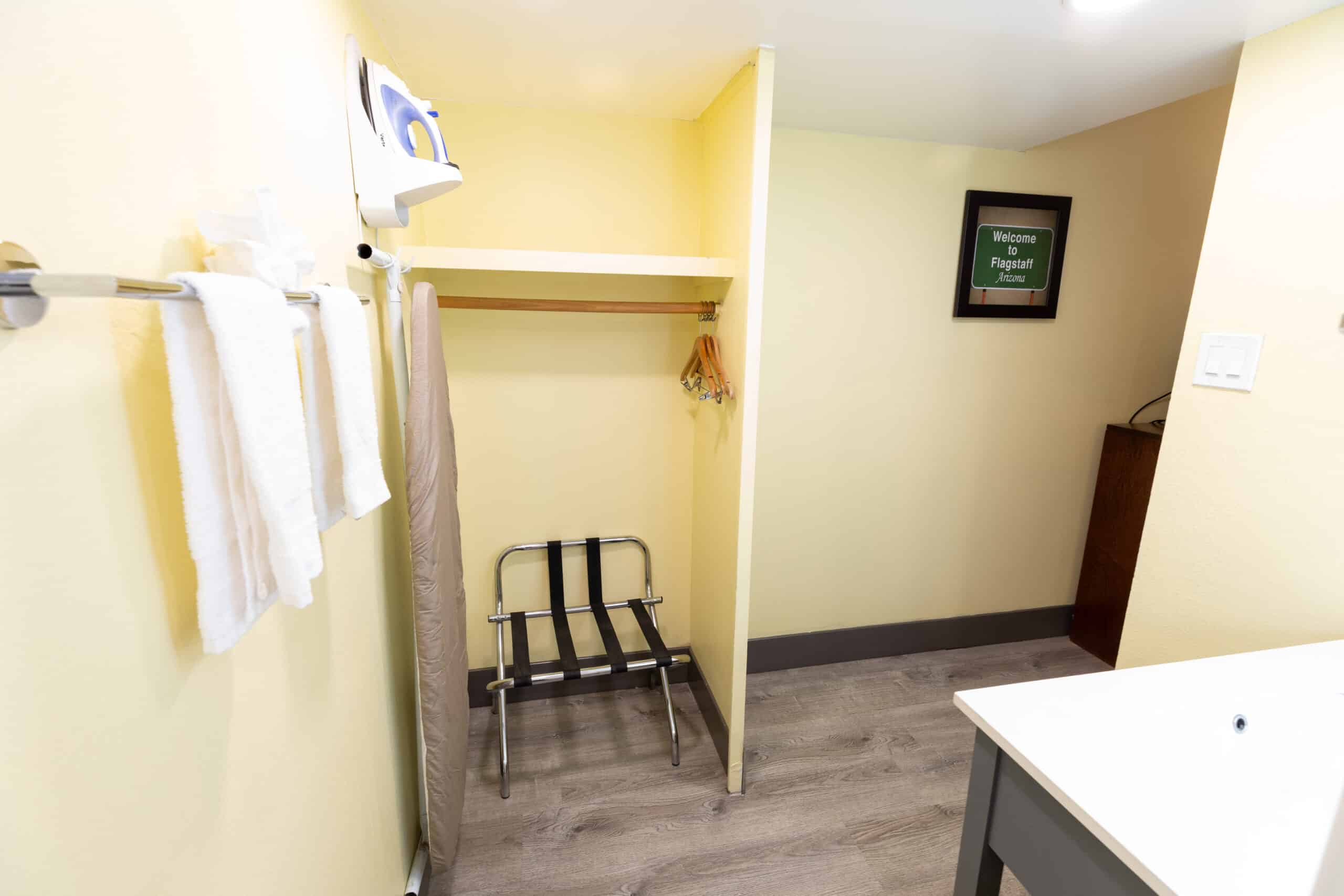 Open closet nook with hanging rack, luggage rack, ironing board, iron, and towel bar.