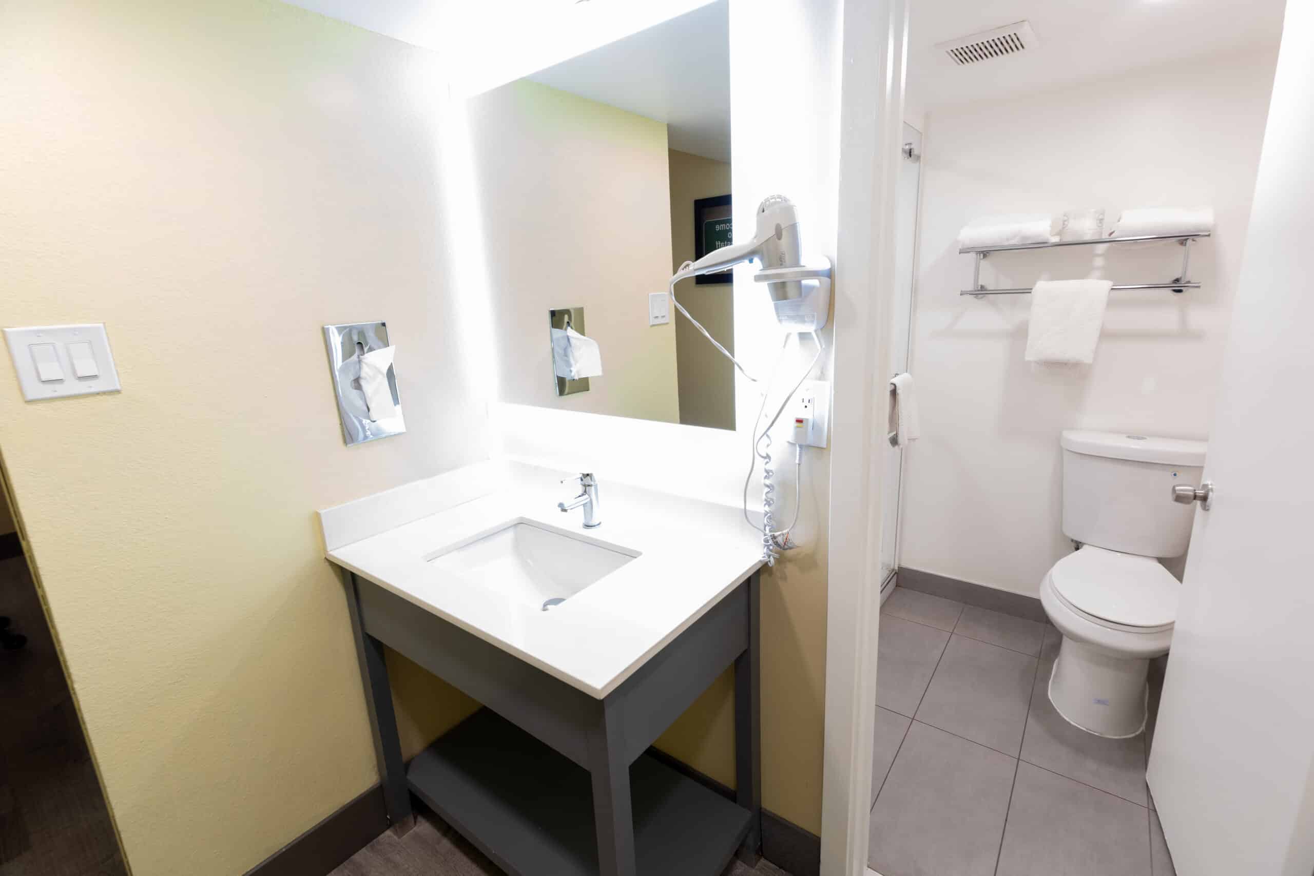 Vanity sink with bright mirror and hair dryer, leading into bathroom with toilet and towel shelf.