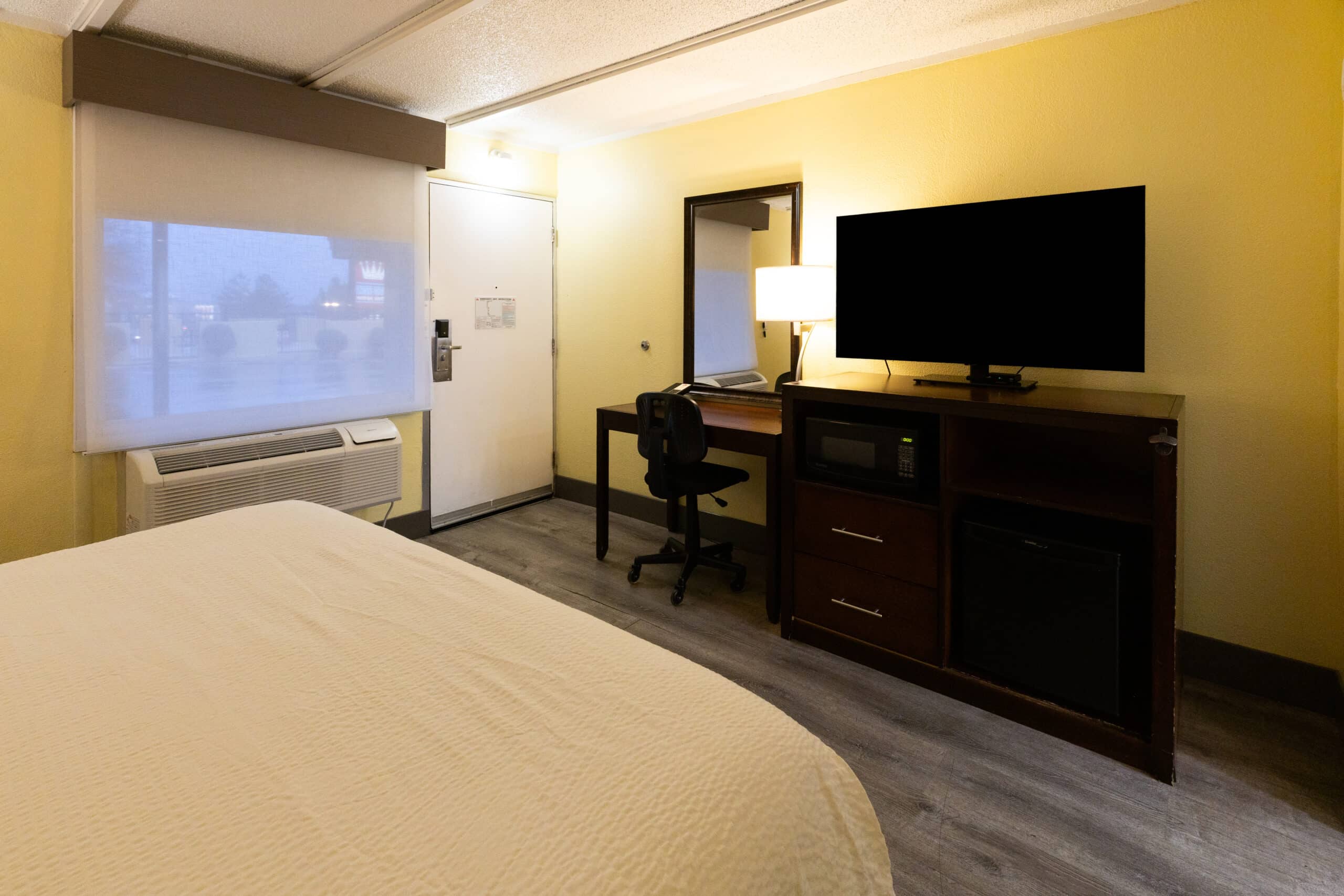 Guest room workspace with desk and chair, TV on cabinet with microwave and mini fridge, and a window shade above the air-conditioning unit.