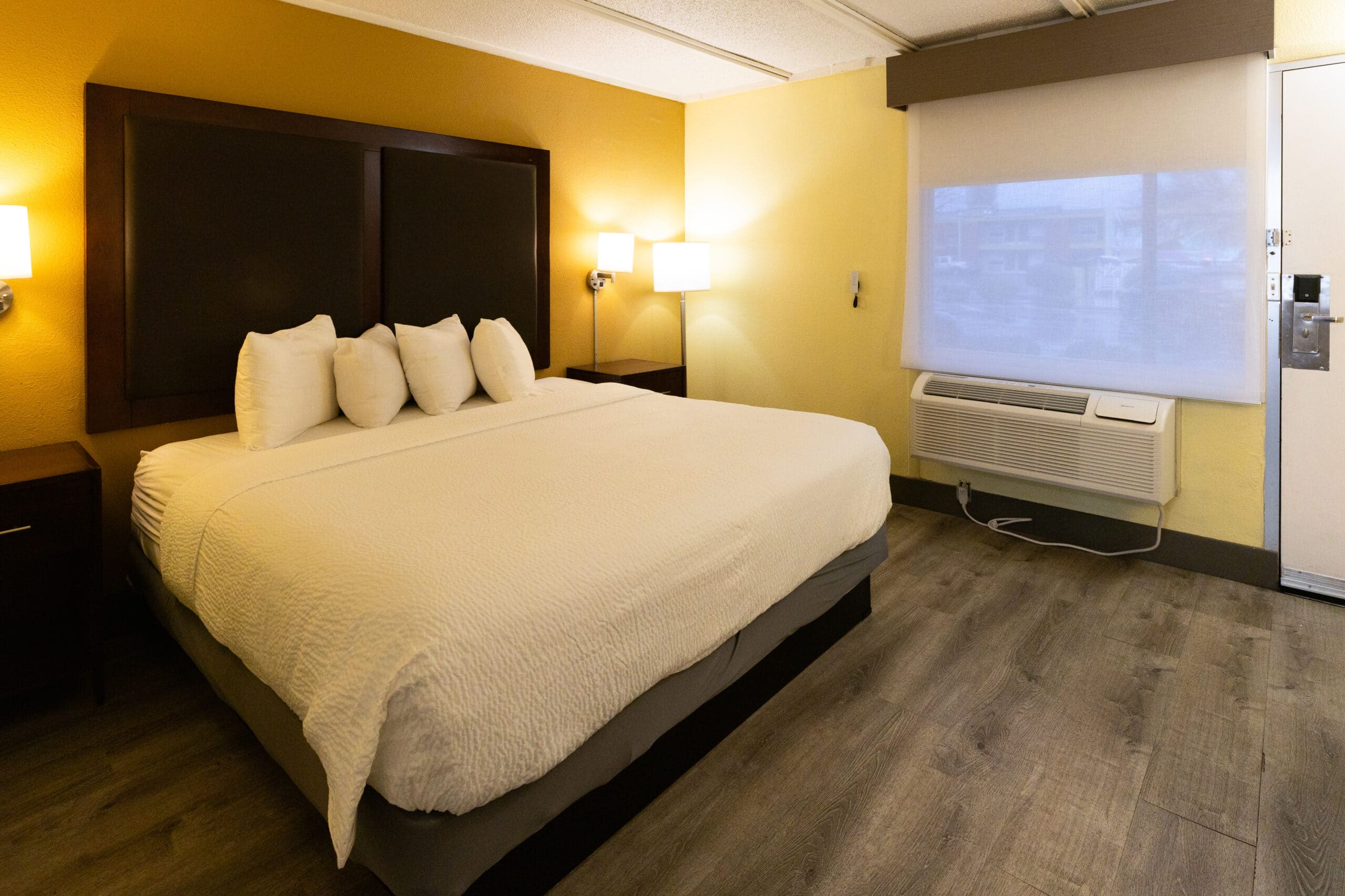 King bed beside a window with roller shade and in-room air-conditioning unit, with bedside lighting and wood-look flooring.