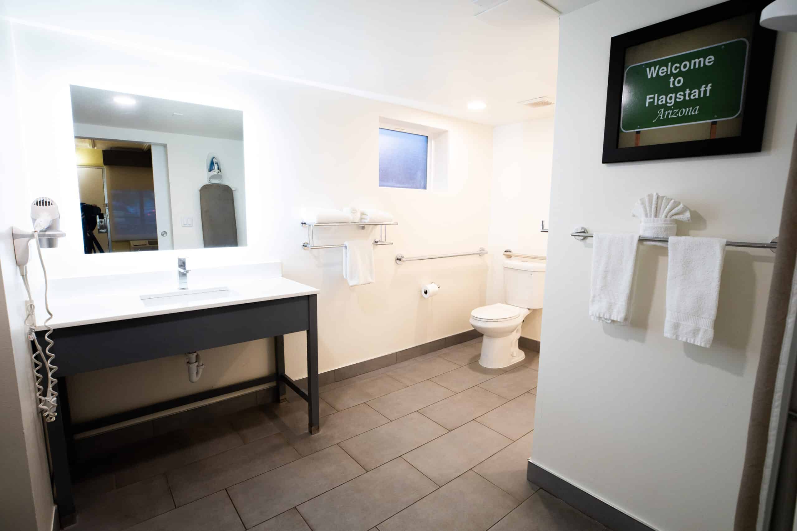 Accessible bathroom with wide vanity, large illuminated mirror, toilet, grab bars, and tiled flooring.