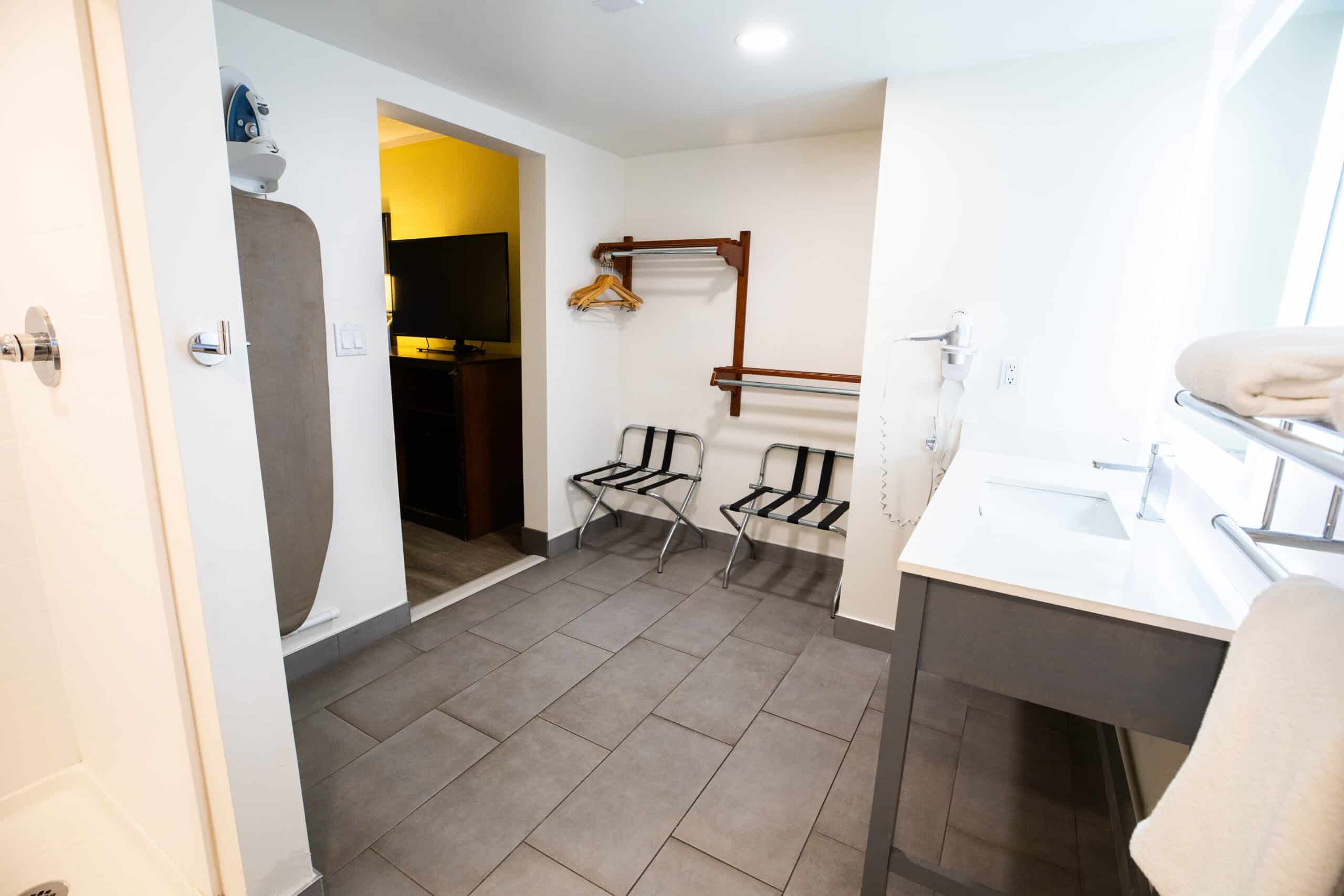 Vanity and dressing area with open closet, hangers, luggage rack, ironing board, and sink counter next to the guest room.