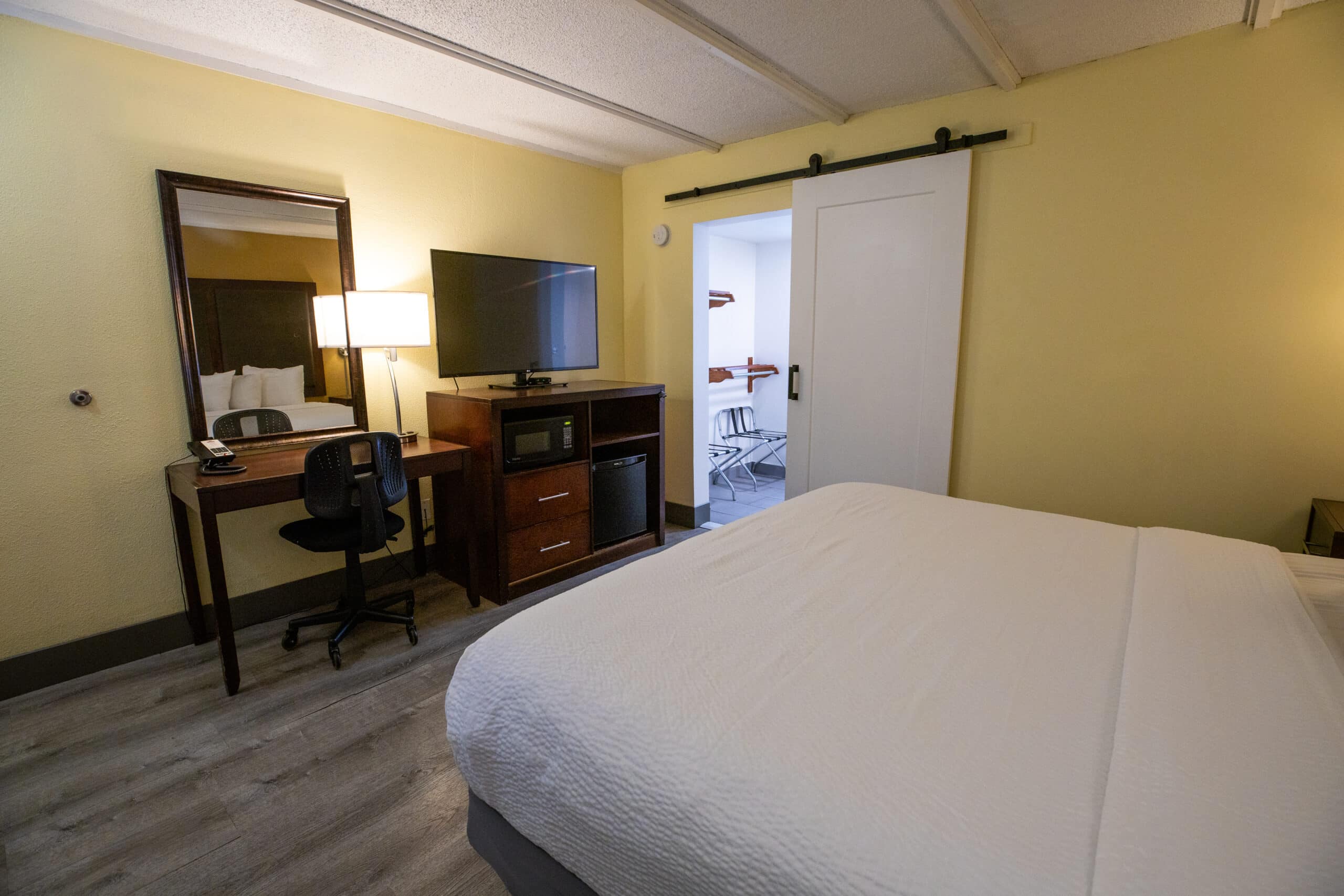 View from the bed toward a TV on a cabinet with microwave and mini fridge, a desk workspace, and a sliding barn door to the vanity area.