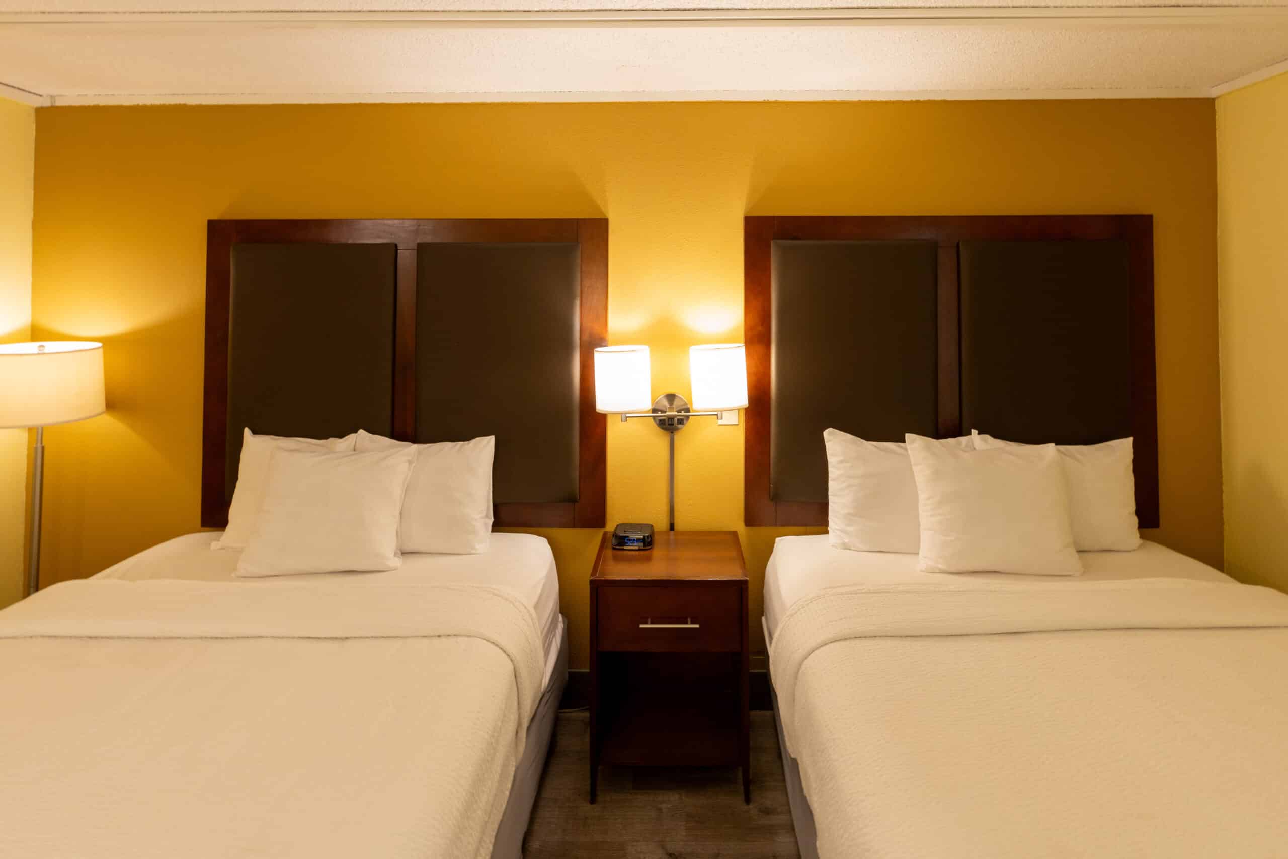 Two queen beds in Kings House guest room with nightstand, wall lamps, and warm accent wall